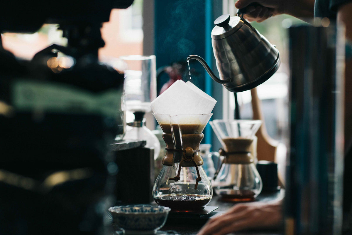 A gooseneck kettle pouring into a pour-over brewer set over glass carafes on a bar.