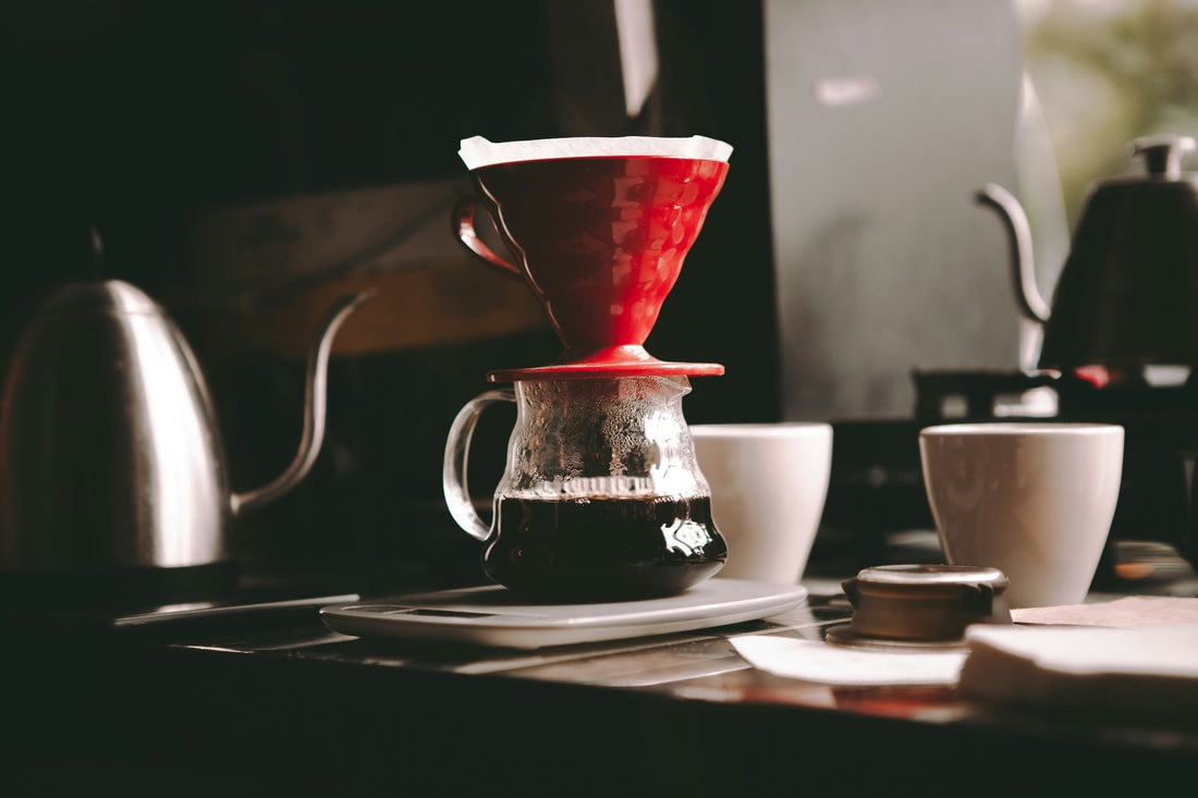 A red pour-over dripper sitting on a glass carafe beside a gooseneck kettle.