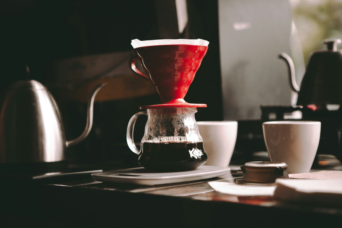 A red pour-over dripper sitting on a glass carafe beside a gooseneck kettle.